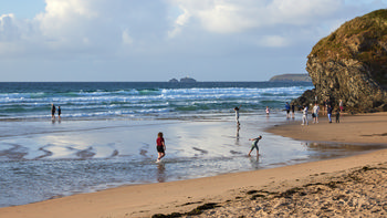 Gwithian beach This landscape photograph captures Gwithian beach in Cornwall, England, during a summer evening. The image shows groups of people walking and playing along the shoreline, with gentle waves rolling in from the Atlantic Ocean. A prominent rocky outcrop is visible to the right side of the frame, adding interest to the composition. In the distance, Godrevy Lighthouse, a well-known landmark in Cornwall, can be seen on an island just offshore. The scene is characterised by the warm light of evening, creating reflected patterns on the wet sand and highlighting the coastal scenery of the United Kingdom.